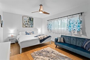 Une chambre moderne avec un lit, un canapé bleu, un ventilateur de plafond et une fenêtre ornée de rideaux à motifs floraux.