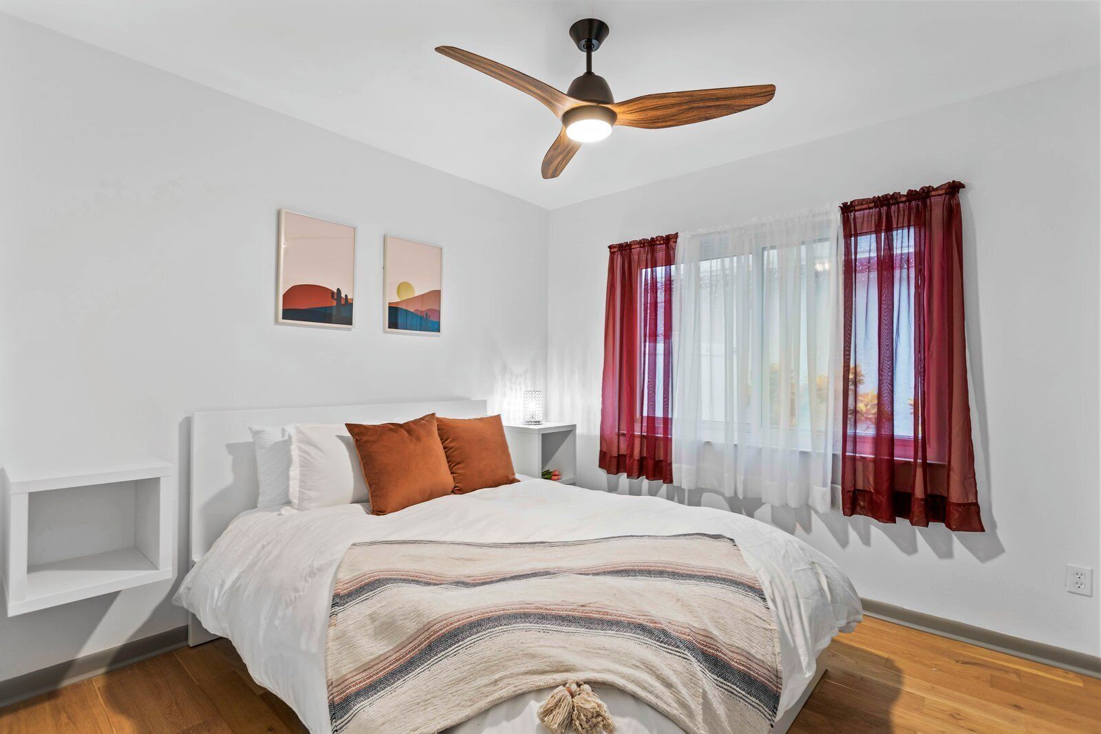 A bedroom with a bed, a ceiling fan, two wall-mounted artworks, and sheer red curtains.
