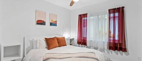 A bedroom with a bed, a ceiling fan, two wall-mounted artworks, and sheer red curtains.