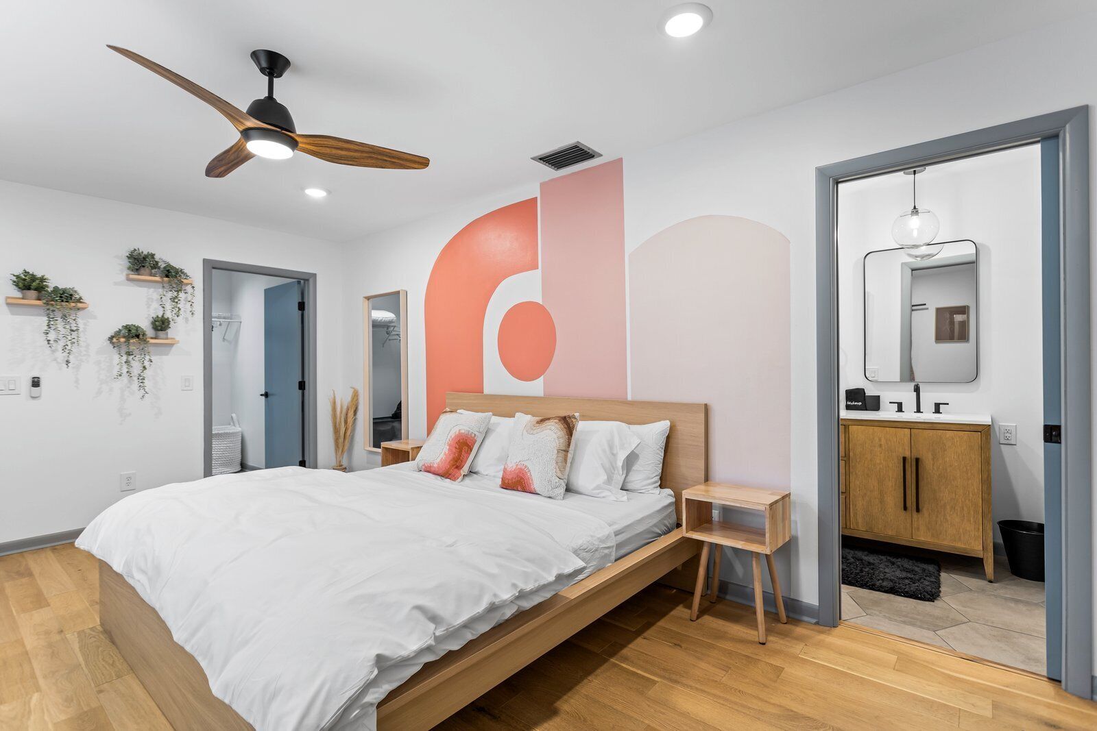 Un dormitorio moderno con una cama de madera, ventilador de techo, un espejo grande y un baño minimalista.