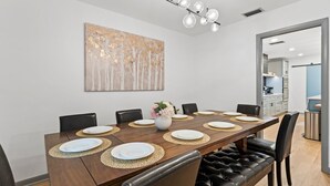 A dining area with a wooden table set for six, surrounded by black leather chairs, a modern chandelier, and a large abstract painting on the wall. - Luxury Oasis w/ Private Pool - 5-Minutes to Beach (Seminole)