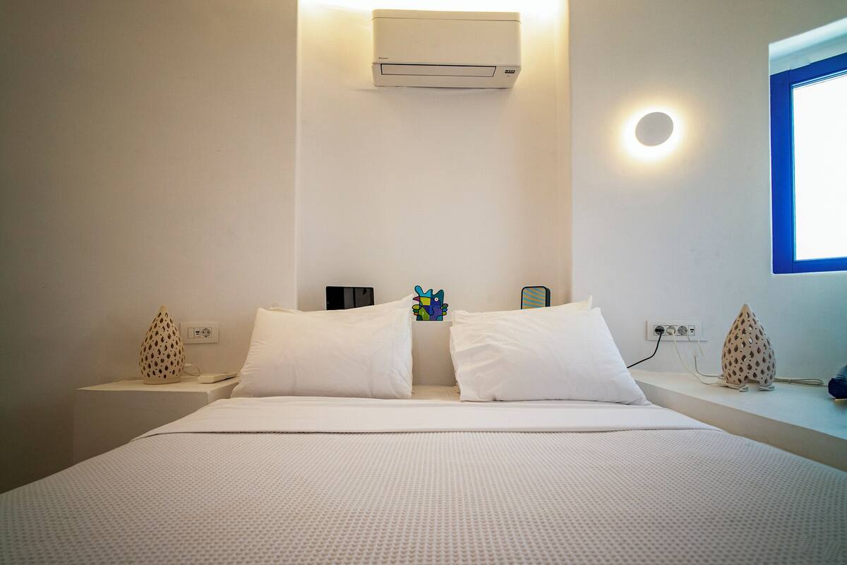 A hotel room with a bed, two white pillows, a wall-mounted air conditioner, a bedside table with a lamp, and a window with a view.