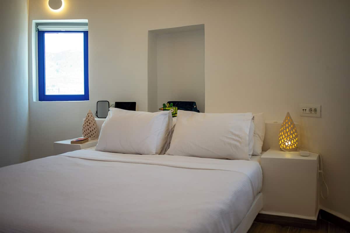 A neatly made bed with white linens, a bedside table with a lamp, and a window with blue blinds.