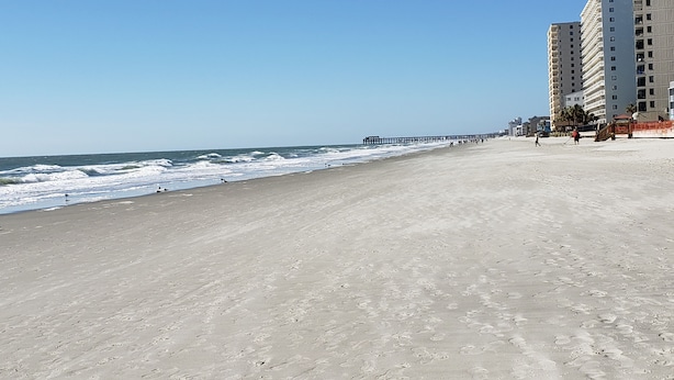 Een strand met een lange steiger die zich uitstrekt over de oceaan, een zandstrand en een hoog gebouw met meerdere balkons aan de rechterkant.