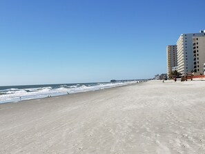 Uma praia com um longo cais que se estende para o oceano, uma faixa de areia e um edifício alto com várias varandas à direita.