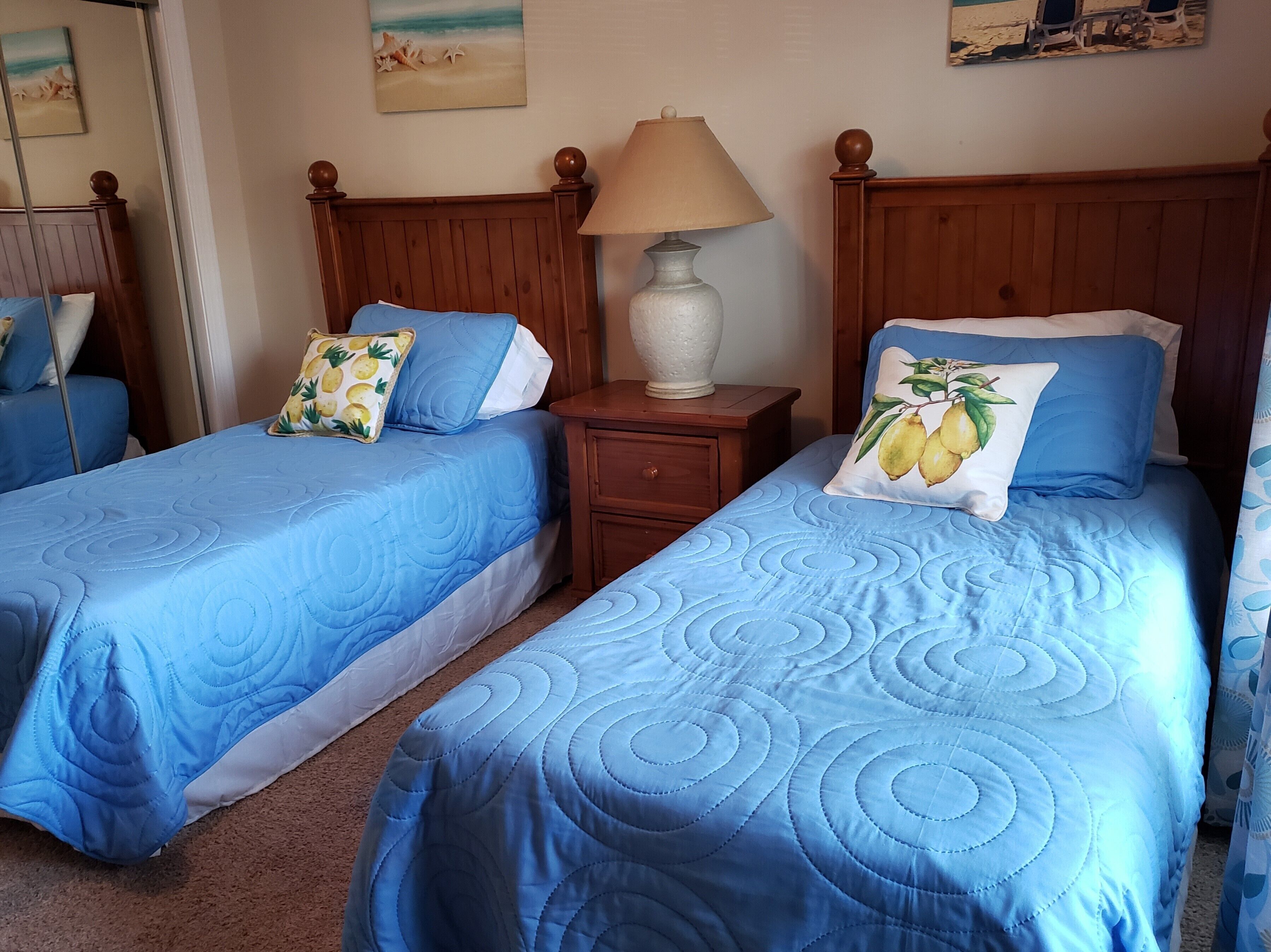 Ein Hotelzimmer mit zwei Betten, jeweils mit blauer Bettwäsche und dekorativen Kissen, einem Holzkopfende, einem Nachttisch mit Lampe und Wandbildern mit Strandmotiven.