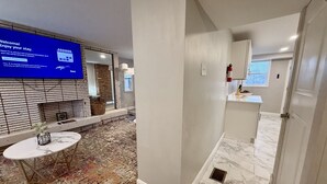 Un couloir d’hôtel avec un téléviseur à écran plat affichant un message de bienvenue, une petite table ronde avec un vase, un tapis à motifs et un coin cuisine avec une fenêtre.