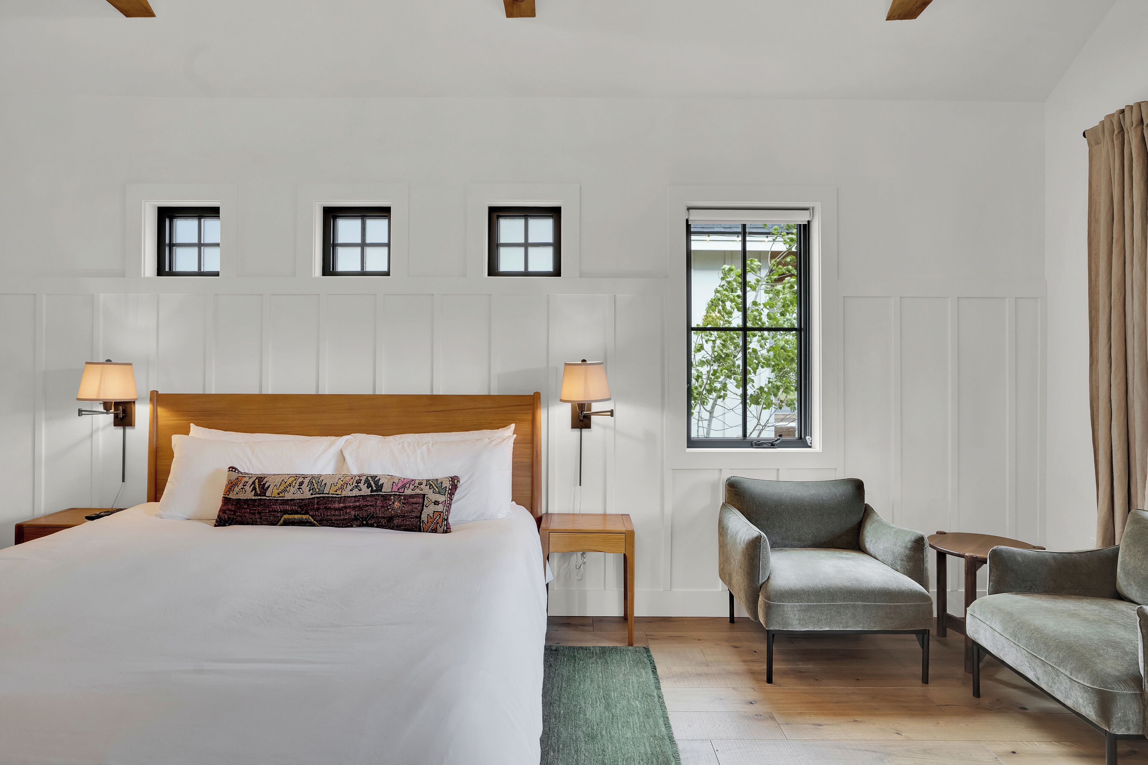 A hotel room with a large bed, two bedside tables with lamps, a chair, a small table, and a window with a view of trees.