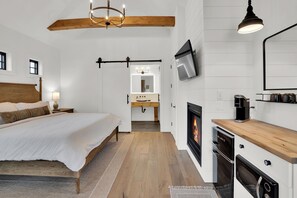 A bedroom with a large bed, a wooden ceiling, a TV, a fireplace, and a bathroom with a mirror and sink.