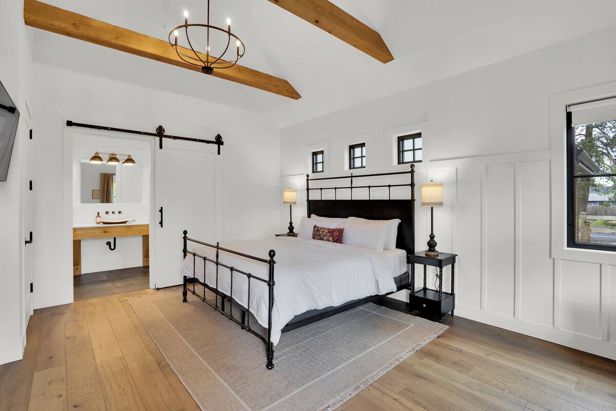 A bedroom with a wooden floor, a bed with white bedding, a nightstand with a lamp, and a large window.
