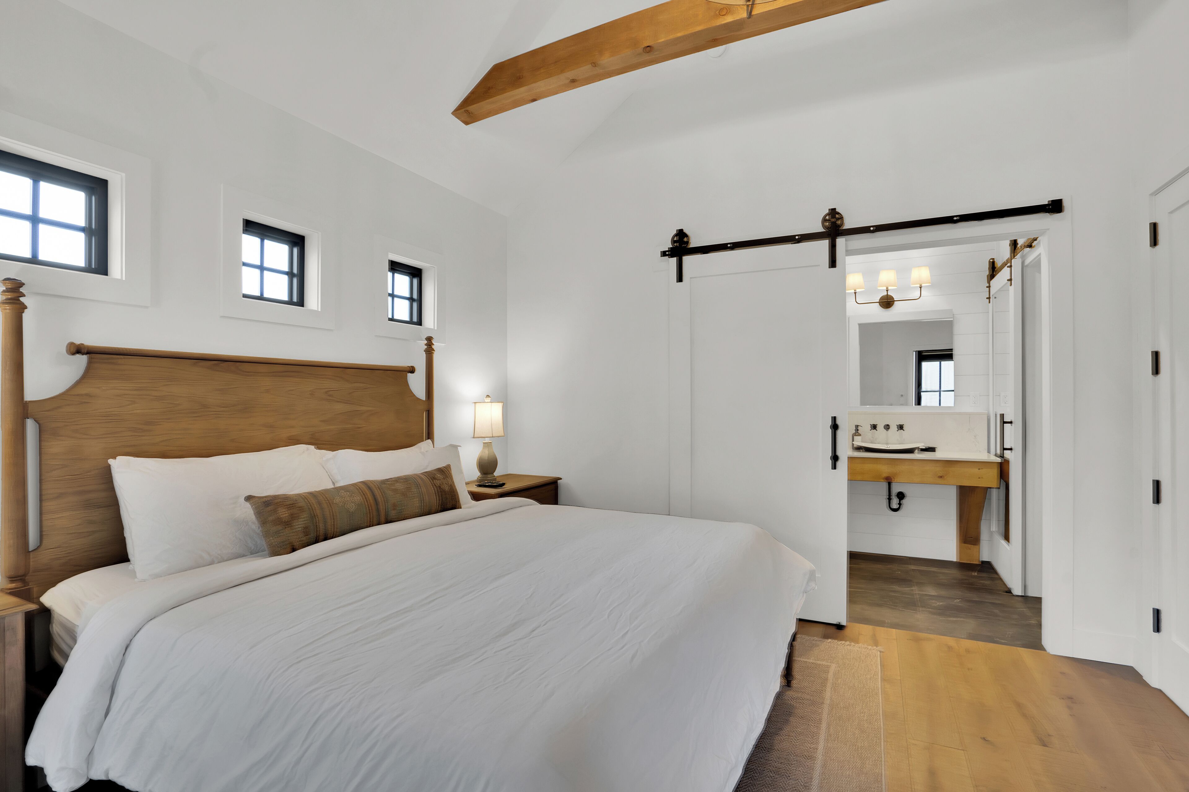 A bedroom with a large bed, wooden headboard, bedside tables with lamps, a wooden ceiling beam, and a view into the bathroom with a sink and mirror.