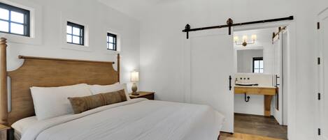 A bedroom with a large bed, wooden headboard, bedside tables with lamps, a wooden ceiling beam, and a view into the bathroom with a sink and mirror.