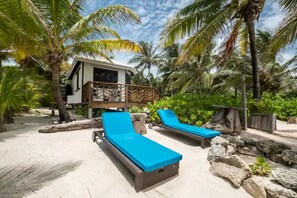 A beachside resort with two sun loungers, a wooden deck, and a small building surrounded by palm trees.