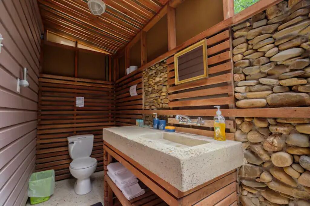 A compact wooden cabin bathroom with a stone wall, a wooden bench, a toilet, and a sink with toiletries.