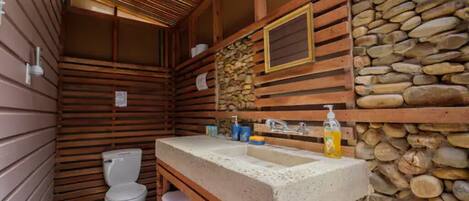 A compact wooden cabin bathroom with a stone wall, a wooden bench, a toilet, and a sink with toiletries.