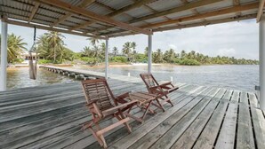 A wooden deck with two lounge chairs overlooking a body of water, surrounded by palm trees and a dock.