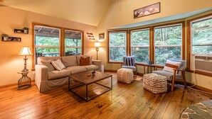 Una sala de estar acogedora con un sofá, un reposapiés y un sillón, pisos de madera, ventanas amplias con vistas a árboles y un ventilador de techo.