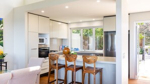 A modern kitchen with white cabinetry, a built-in microwave, and a stainless steel refrigerator. There is a dining area with a wooden table and chairs, and a window providing a view of the outdoors. - 154 Mitchell Parade - Ocean views from every room (Mollymook Beach)