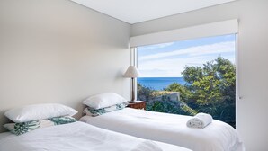 A bedroom with two beds, a nightstand, a lamp, and a view of the sea. - 154 Mitchell Parade - Ocean views from every room (Mollymook Beach)