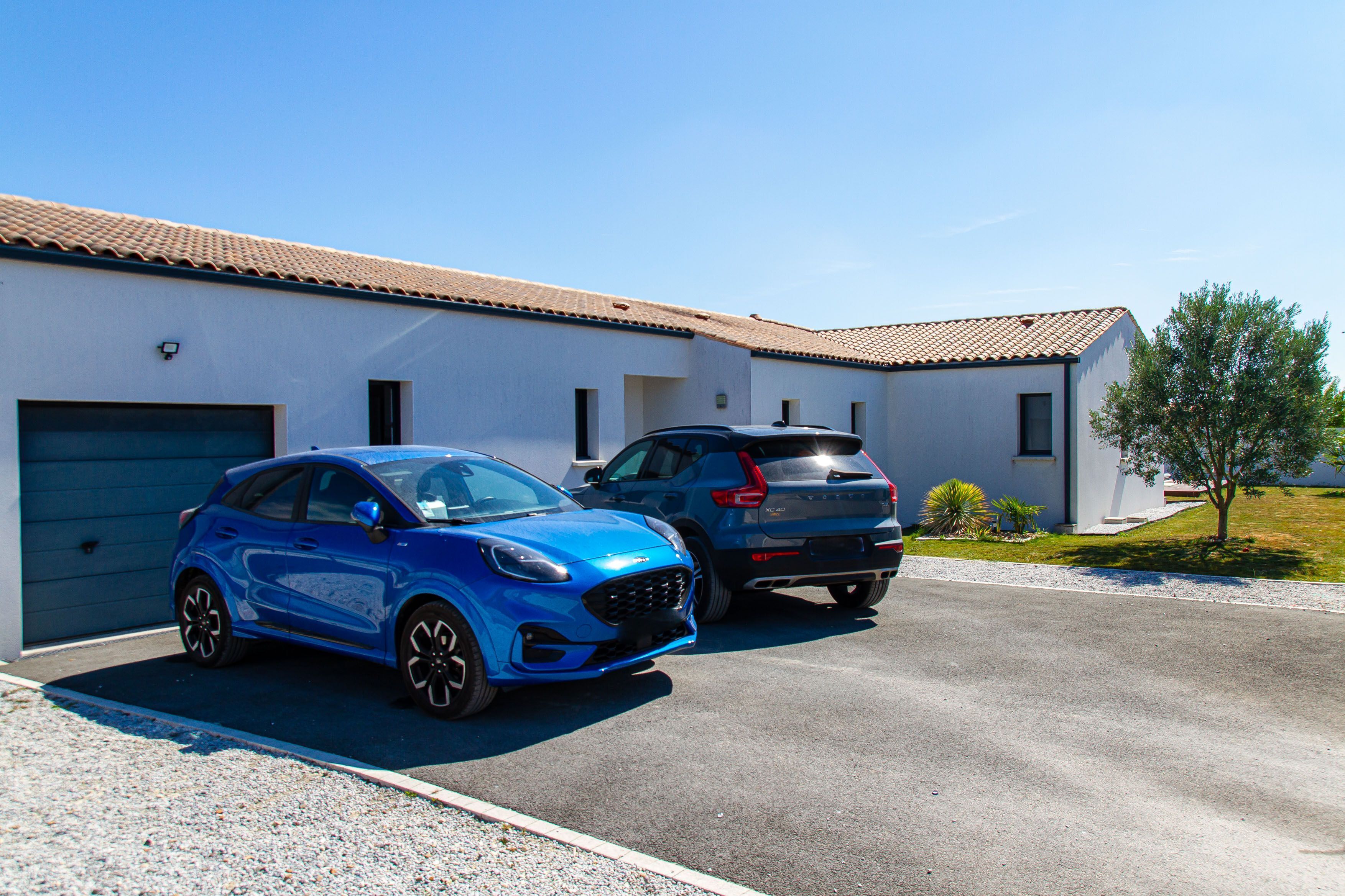 Deux voitures sont garées devant un immeuble dont le toit est recouvert de tuiles et qui possède un garage.