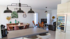 Eine moderne Küche mit Kochinsel, Pendelleuchten, einer Essecke mit Stühlen und einem Kühlschrank.