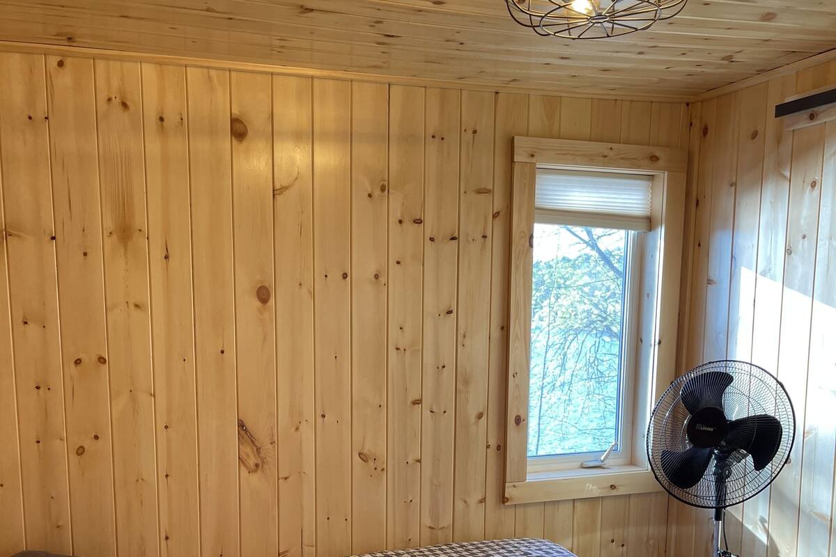 Una habitación con paredes de madera, una cama con colcha estampada, un ventilador de pie y una ventana con persianas.