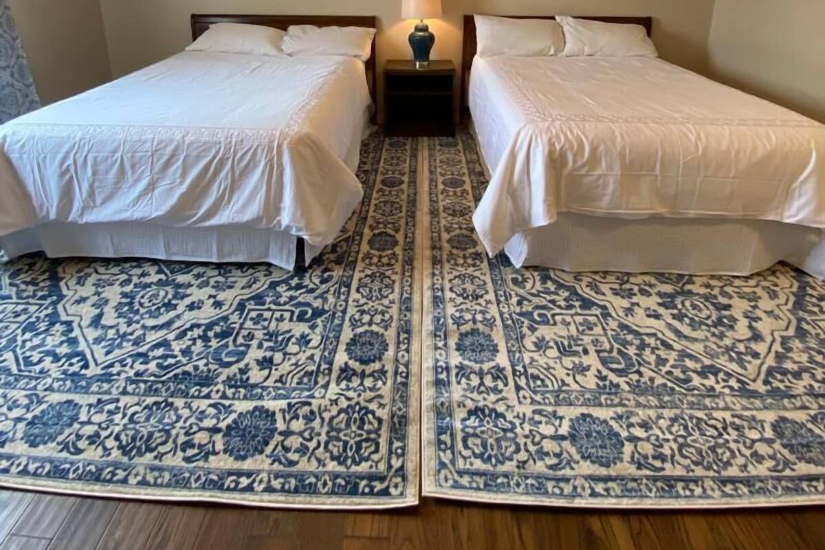 Una habitación con dos camas, un tapete estampado, un ventilador de techo y cuadros enmarcados en la pared.