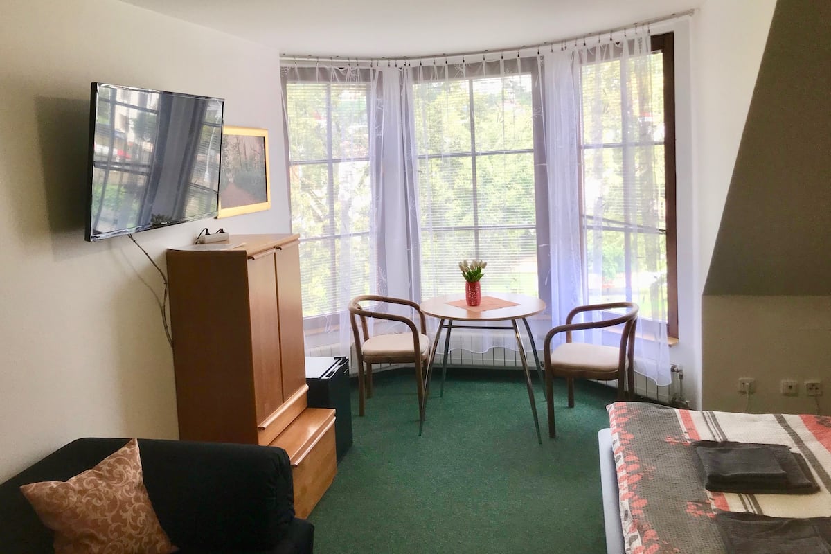 A room with a bed, a TV mounted on the wall, a small table with chairs, and large windows with curtains.