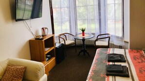 A small, cozy room with a sofa, a table with chairs, a TV mounted on the wall, and large windows with sheer curtains. - Penzion JANY (Luhacovice)