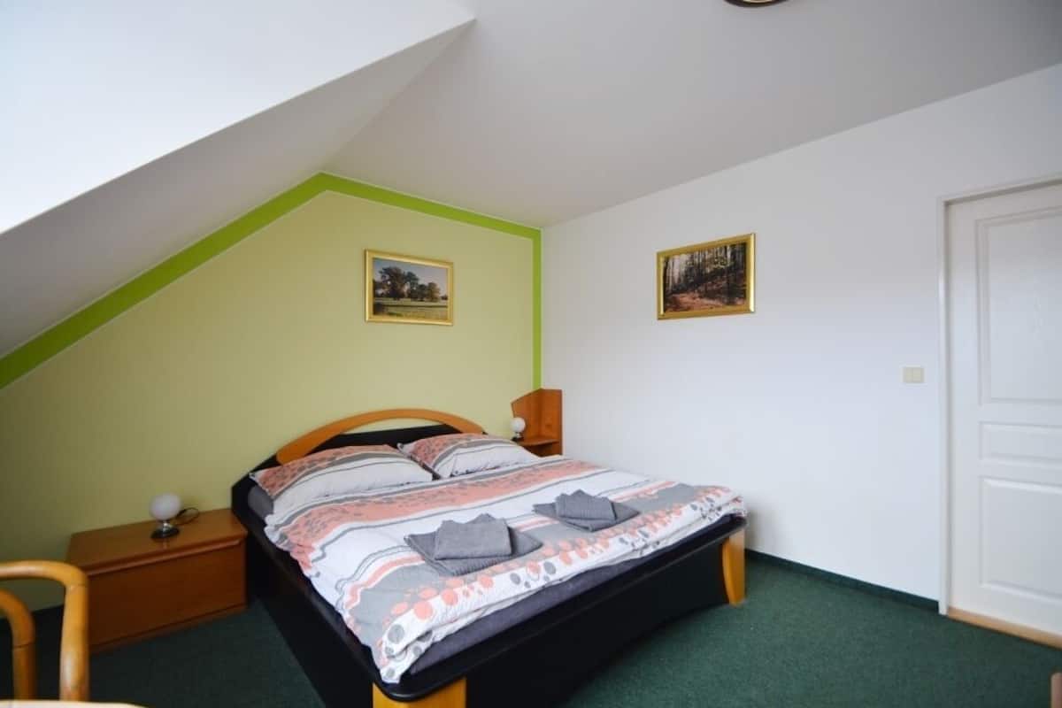 A bedroom with a sloped ceiling, a bed with a patterned bedspread, two framed pictures on the wall, a bedside table with a lamp, and a chair.
