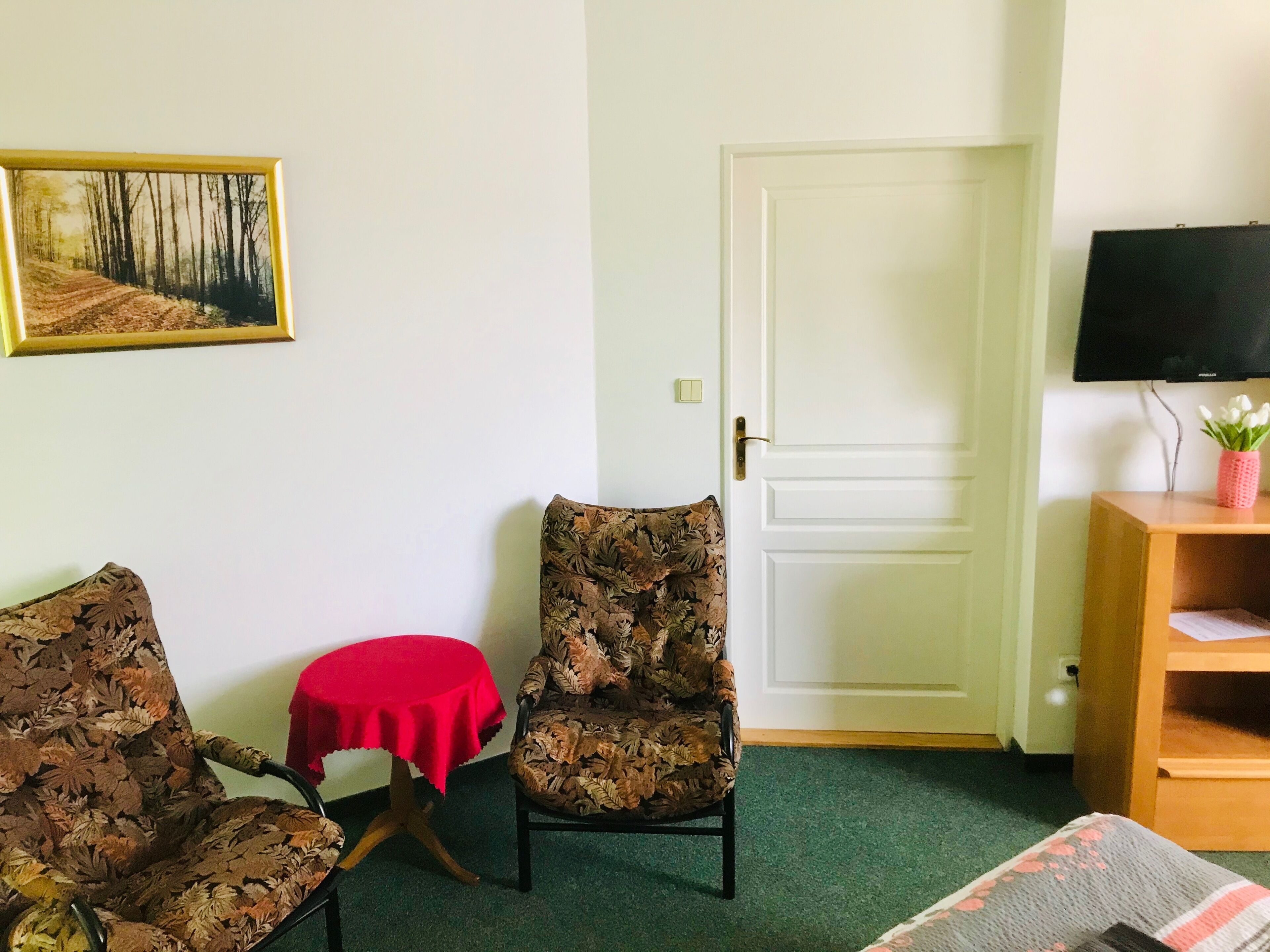 A room with two patterned chairs, a small red table, a television on a wooden stand, and a door.