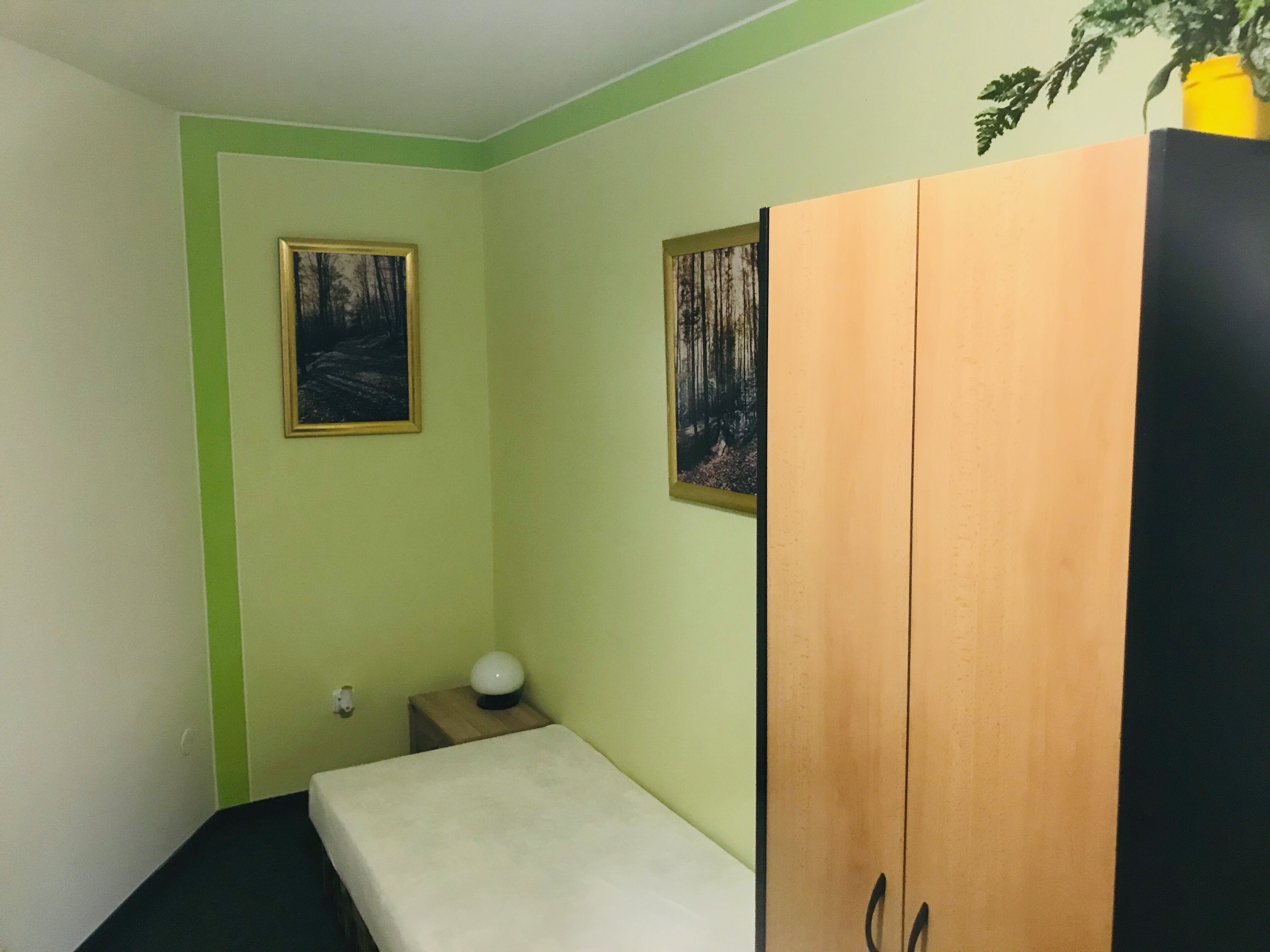 A small, single-bed room with a wooden wardrobe, a bedside table with a lamp, and two framed pictures on the wall.