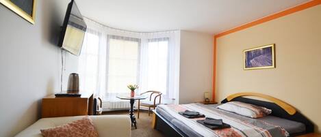 A hotel room with a bed, a sofa, a small table with a plant, a chair, and a television mounted on the wall.