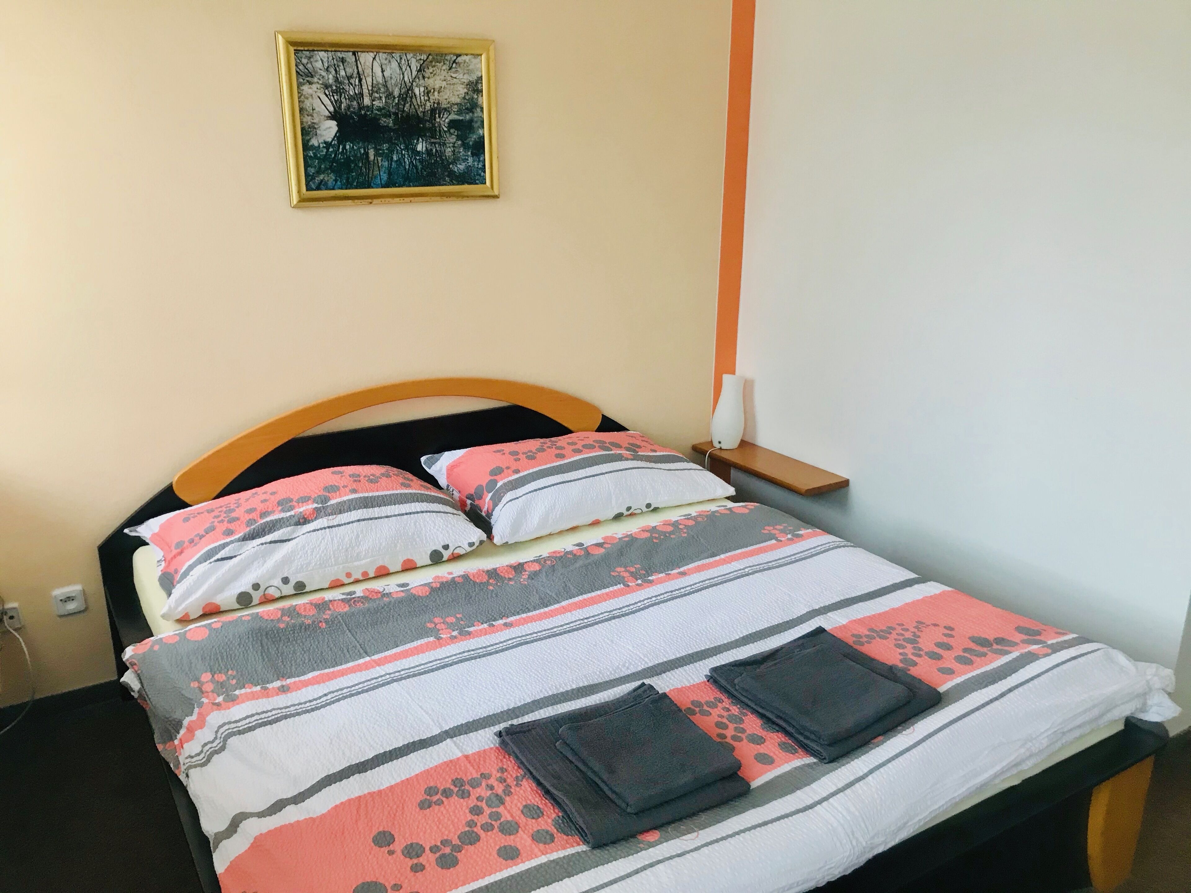 A bed with a striped bedspread, pillows, and folded towels. There is a nightstand with a white lamp and a small shelf. A framed picture hangs on the wall above the bed.