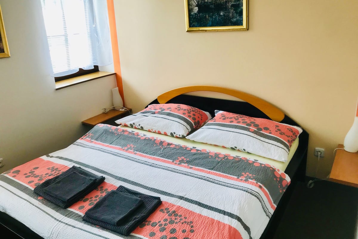 A neatly made bed with striped bedding and two pillows, a bedside table with a lamp, and a window with blinds.