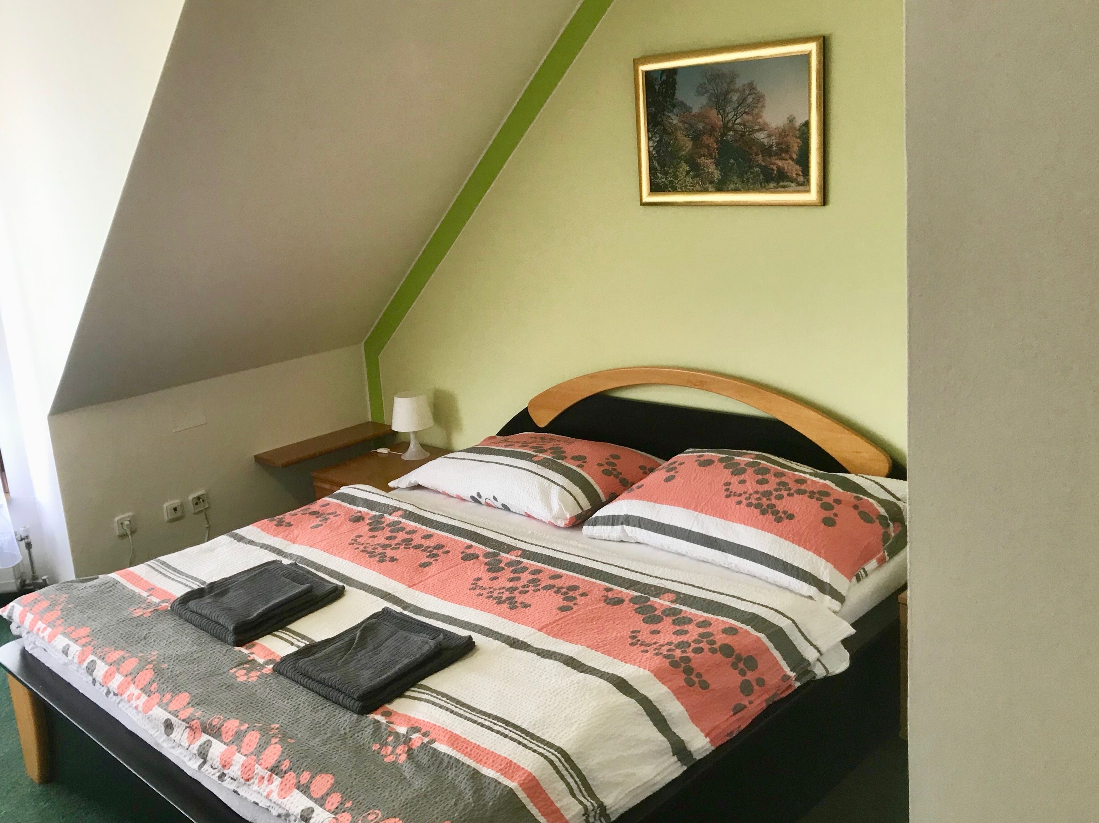 A bedroom with a sloped ceiling, a bed with a floral-patterned bedspread, two pillows, a bedside lamp, and a framed picture on the wall.