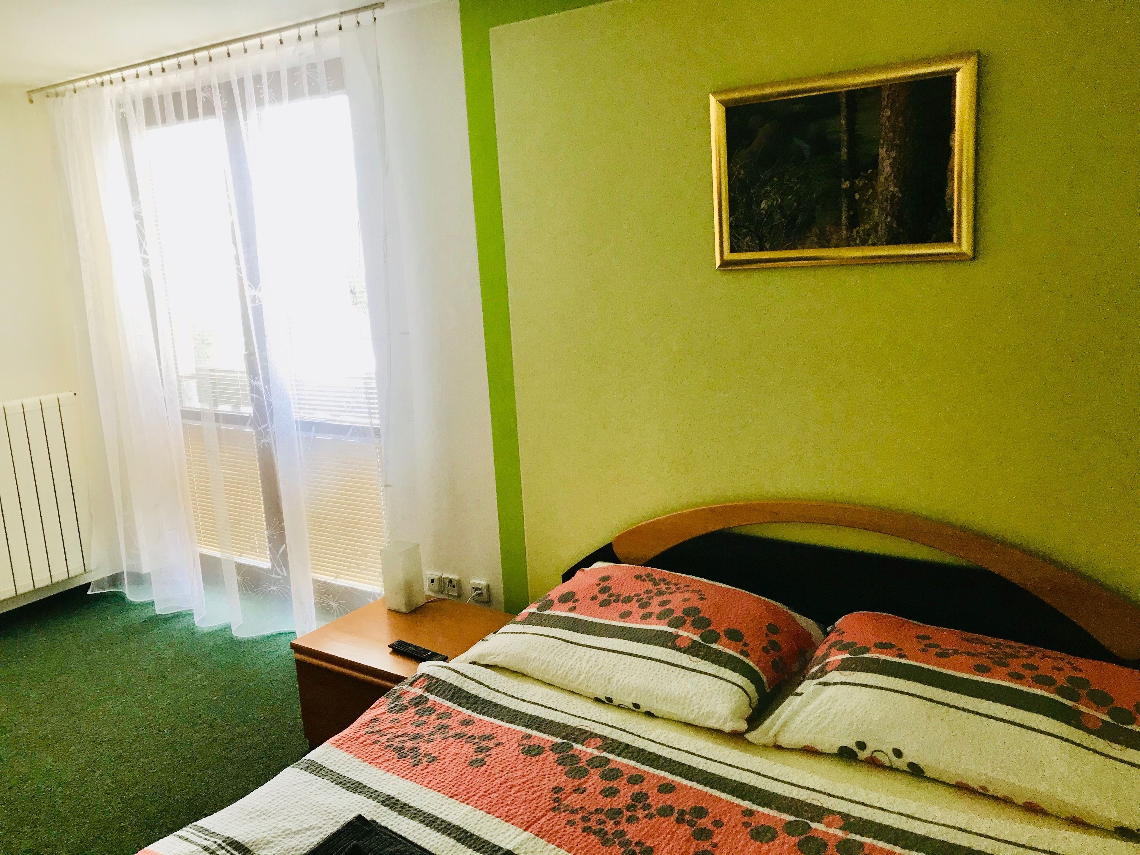 A bedroom with a bed, a nightstand, a window with sheer curtains, a radiator, and a green wall with a framed picture.
