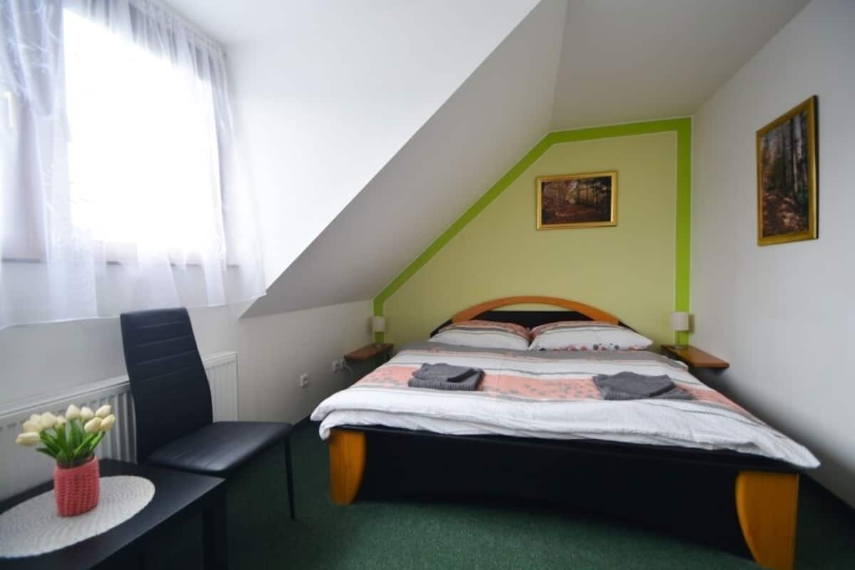 A bedroom with a bed, a chair, a small table with a vase of flowers, and a window with curtains.