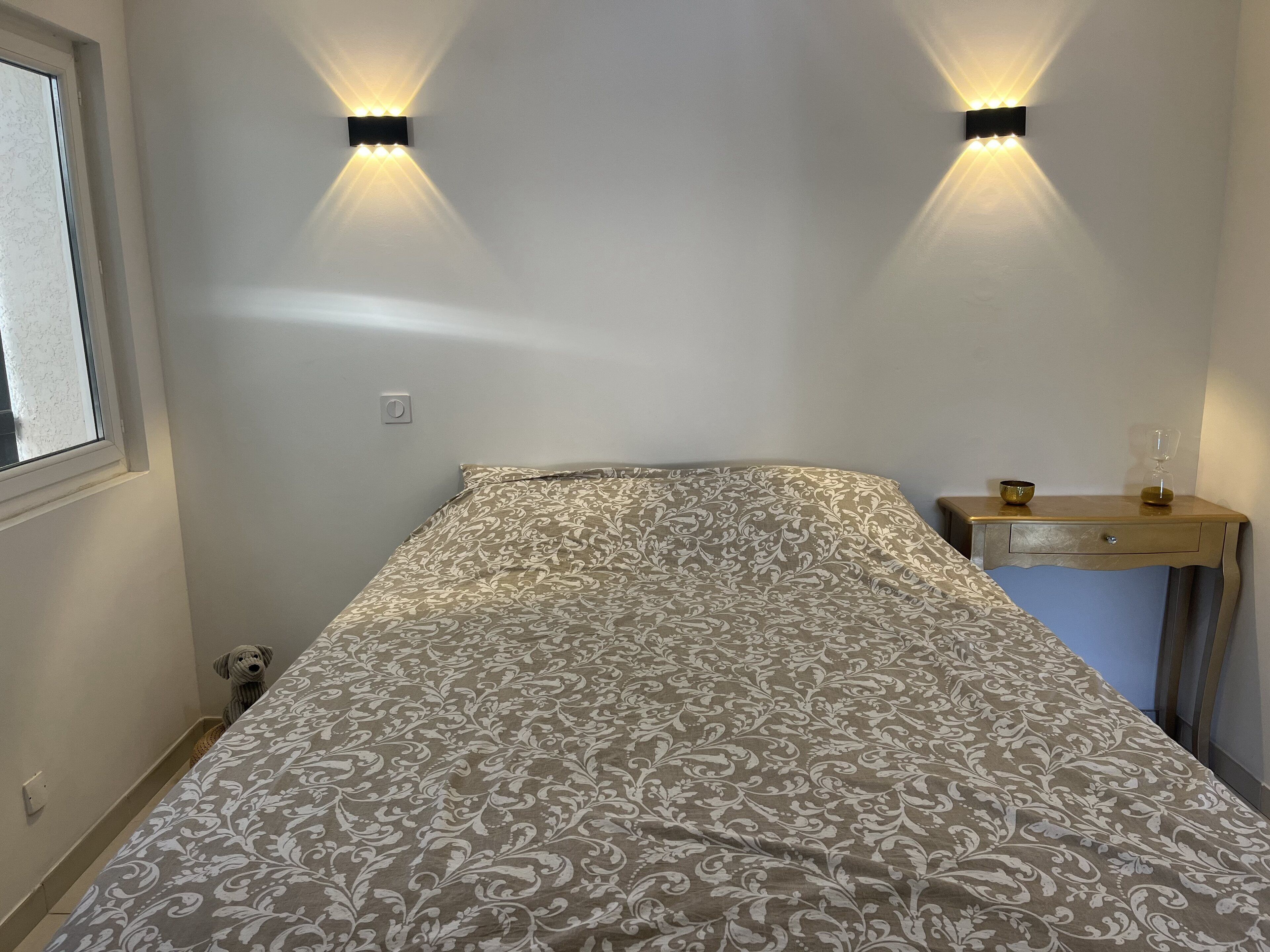 Un dormitorio con una colcha estampada, una mesita de noche con una lámpara y dos luces en la pared.