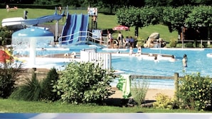 Une piscine avec des toboggans et un espace aquatique, le tout entouré d’arbres et de pelouses.