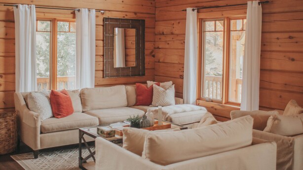 A cozy log cabin interior with a large wooden ceiling, a white sofa set, and a window with curtains.