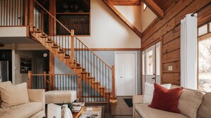 A spacious living room with a wooden staircase, a sofa, a coffee table with books and decorative items, and a view of the outside.