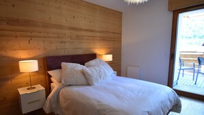 A bedroom with a wooden headboard, a bed with white bedding, two bedside lamps, and a view of an outdoor area through a glass door. - L'Étendard appartement 5 (Vaujany)