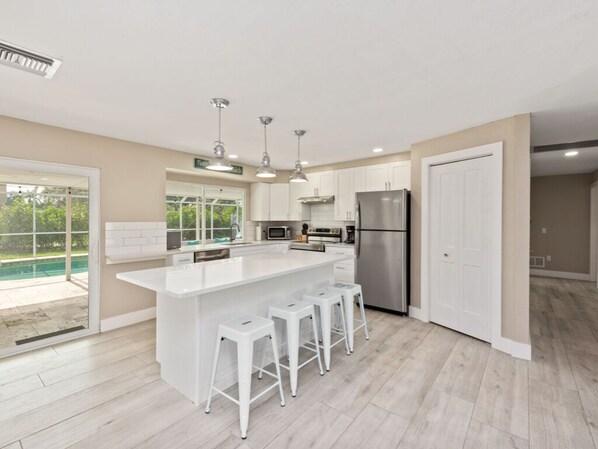 A modern kitchen with a central island, white bar stools, stainless steel appliances, and a view of the outdoors. - Mackle Park Family Getaway (Marco Island)