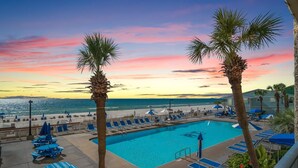 Zona de piscina con sillones reclinables y sombrillas, con vistas a una playa y al océano al atardecer.