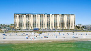 Un hotel grande con varios pisos y balcones, una playa con tumbonas y sombrillas, y un cielo azul despejado.