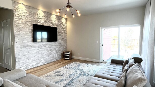 A spacious living room with a large sectional sofa, a flat-screen TV mounted on a textured wall, a contemporary chandelier, and a sliding glass door leading to an outdoor area. - Updated Great Location Beautiful New House (Thornton)