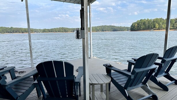 A covered deck with Adirondack chairs and a small table, overlooking a lake with a forested shoreline. - Lakefront Hideaway on Hartwell (Lavonia)