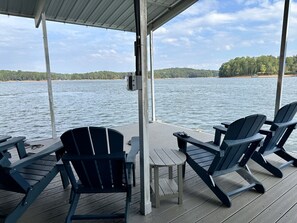 Une terrasse couverte, dotée de chaises Adirondack et d’une petite table, offre une vue sur un lac bordé d’une forêt.