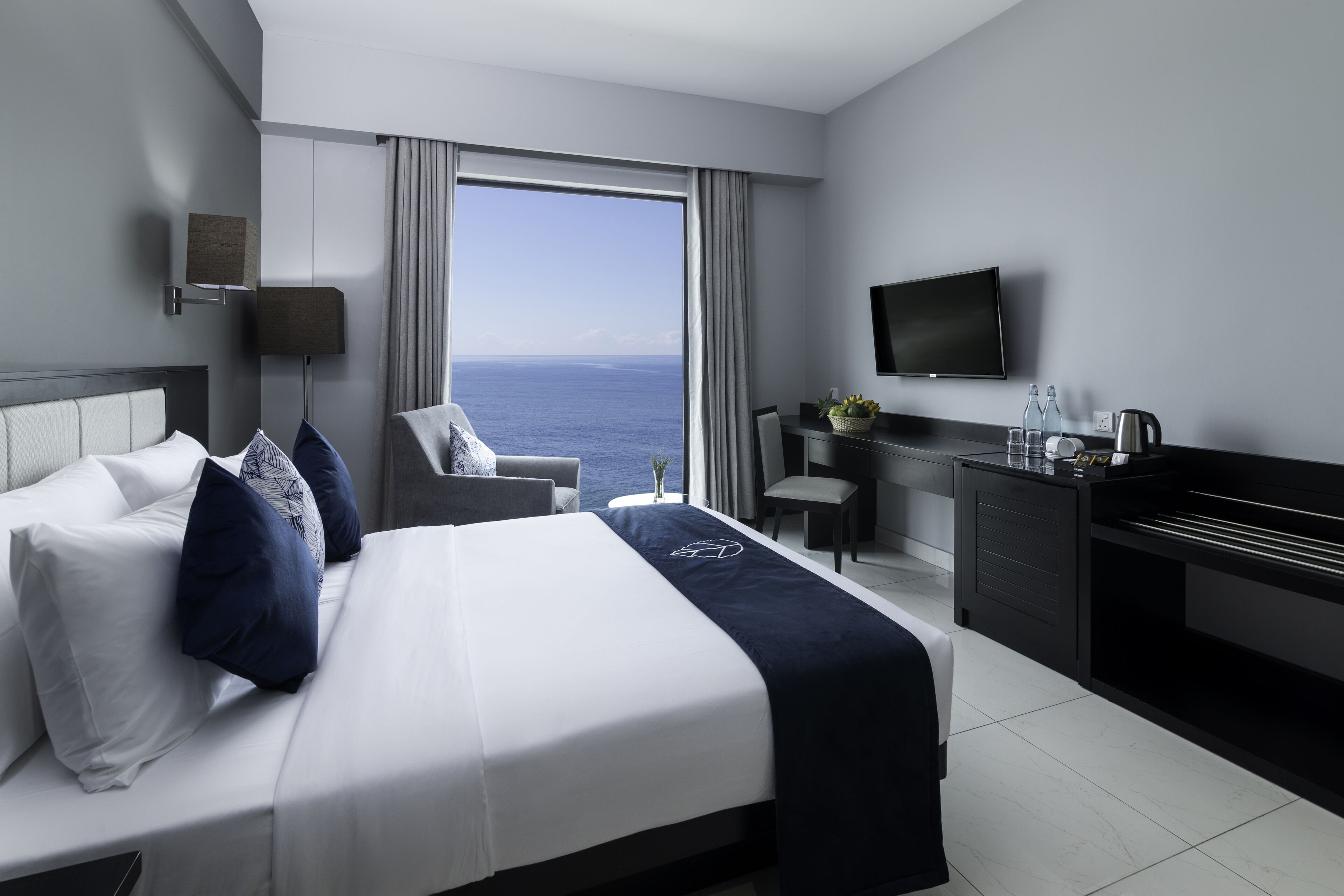 A hotel room with a large bed, a desk, a chair, a television, and a view of the ocean.